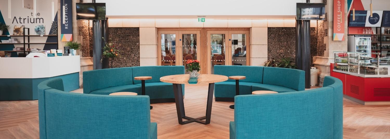 Cushioned turquoise seating arranged in a large circle with small tables on a wooden floor in the Atrium entrance. Glass doors looking into the church sanctuary in the background.